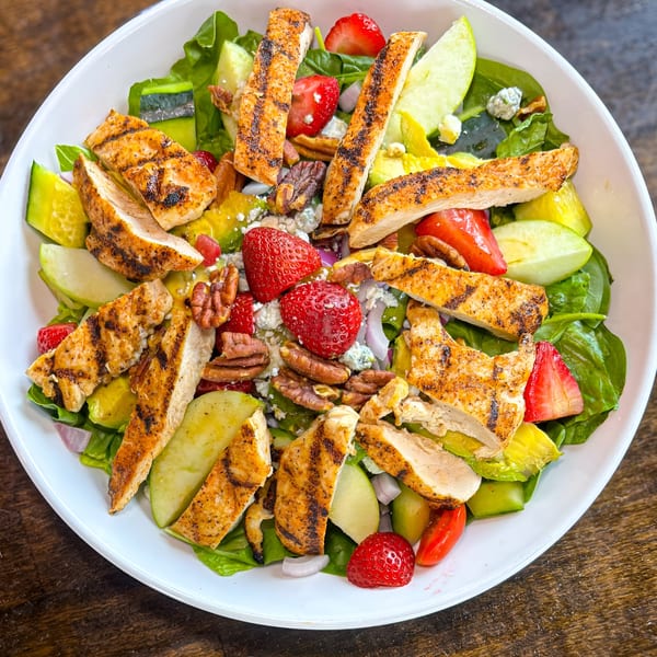 Grilled chicken salad with strawberries, pecans, apples, and blue cheese crumbles in a white bowl.