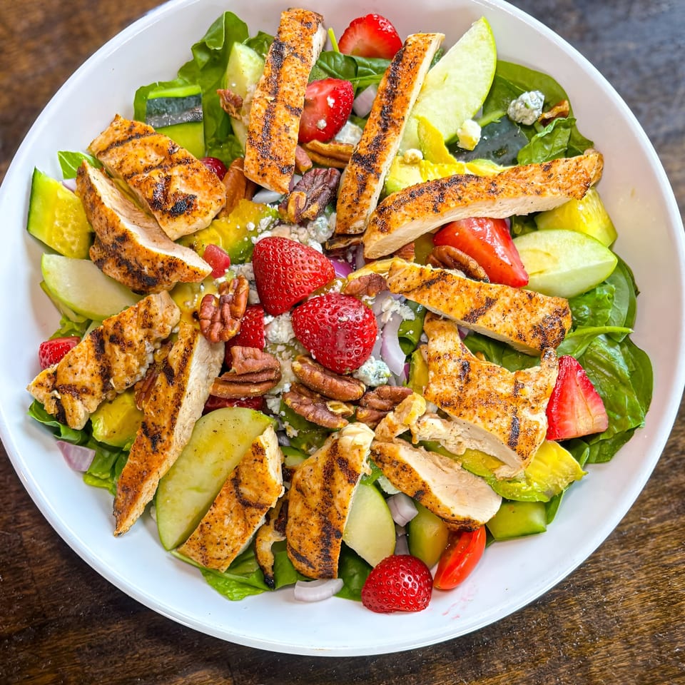Grilled chicken salad with strawberries, pecans, apples, and blue cheese crumbles in a white bowl.
